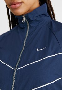 Casaco azul marinho com fecho éclair, colarinho alto, riscas brancas de destaque e um pequeno logótipo da Nike em branco no peito. Feito de material leve.