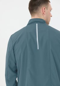 Teal jacka med en slät, matt yta. Har en ståkrage, en subtil grå vertikal stripe på baksidan och skräddarsydda axelsömmar.