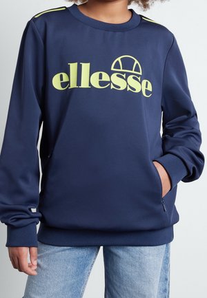 Person trägt einen marineblauen Pullover mit gelbem "ellesse"-Logo und hellblauen Jeans, eine Hand in der Tasche des Pullovers.