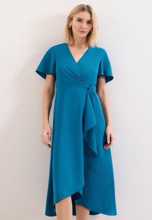 Vrouw met blond haar die een teal-wikkeljurk draagt met zwierige mouwen en een asymmetrische zoom, staand tegen een effen witte achtergrond.