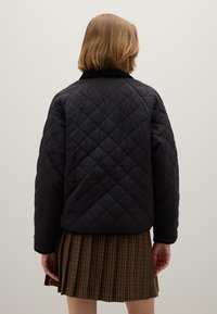 Giacca nera trapuntata con colletto rotondo e bordi a coste, caratterizzata da motivi a diamante. Chi la indossa ha una gonna marrone plissettata.