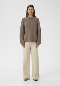 Brauner strukturierter Pullover mit langen Ärmeln, rundem Ausschnitt, kombiniert mit beigen, weiten Hosen und flachen schwarzen Sandalen.