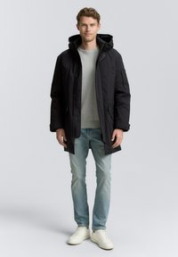 Schwarze Parkajacke mit Kapuze, Frontreißverschluss und Druckknopfverschluss. Verfügt über seitliche Taschen und eine lockere Passform, kombiniert mit hellblauen Jeans und weißen Schuhen.