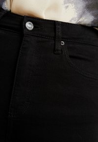 Los jeans de mezclilla negros cuentan con un cierre de botón de metal, costuras discretas en los bolsillos frontales y una textura suave, resaltando elementos de diseño moderno.
