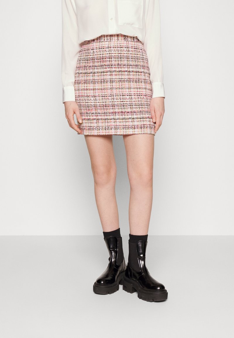 Femme portant une mini-jupe rose à carreaux, un chemisier blanc, des chaussettes noires et des bottines noires en cuir épaisses, debout devant un fond blanc.
