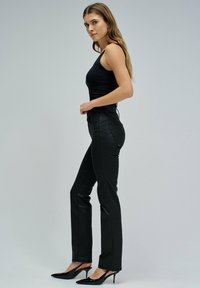 Kobieta z długimi włosami, ubrana w czarny top bez rękawów, czarne spodnie z wysokim stanem i czarne buty na obcasie z odkrytymi piętami, stojąca bokiem.