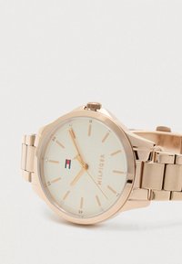 Růžově zlaté hodinky s krémovým ciferníkem, minimálními hodinovými značkami a logem Tommy Hilfiger na levé straně číselníku.