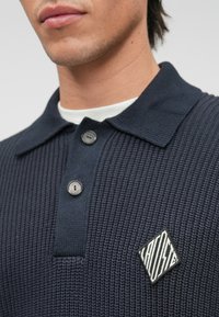 Polo en maille bleu marine avec un motif texturé, un col côtelé, deux boutons et un patch en forme de diamant avec un design noir et blanc.
