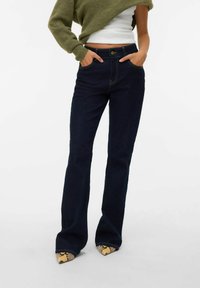 Donkerblauwe flared jeans met gouden stiksels, met een hoge taille en twee zijzakken. Het model draagt puntige hakken met een slangenpatroon.
