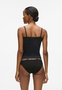 Γυναίκα με μαλλιά σε κότσο που φορά μαύρο τοπ και μαύρα εσώρουχα με την ετικέτα "KARL LAGERFELD" να φαίνεται στο πίσω μέρος, σε άσπρο φόντο.