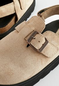 Sandales en daim beige avec une semelle en caoutchouc noir. Présentent une fermeture à boucle et un design découpé sur le devant. Surface texturée pour une meilleure adhérence.