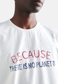 Camiseta blanca de algodón con escote redondo, que presenta un texto desgastado en rojo y azul que dice "PORQUE NO HAY UN PLANETA B."