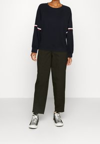 Maglione navy con collo a giro e accenti a righe rosse e bianche, abbinato a pantaloni verde scuro e sneaker alte in bianco e nero.
