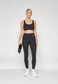 Czarny stanik sportowy i legginsy z wysokim stanem mają miękką tkaninę, dopasowany krój i akcent logo. Modelka trzyma butelkę z wodą.