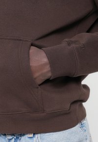Καφέ φούτερ με τσέπη εμπρός και ριμπ μανσέτες, με απαλό ύφασμα. Στο σχέδιο υπάρχει διακριτική λεπτομέρεια με λογότυπο στο μανίκι.