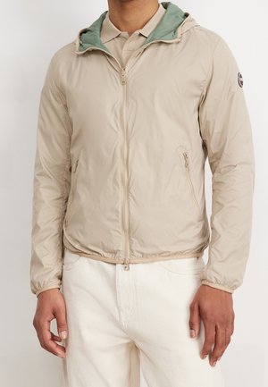 Mann trägt eine beige leichte Jacke mit Reißverschluss und grünem Futter sowie weiße Hose, steht vor einem schlichten Hintergrund.