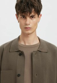 Hnědá pletená košilová bunda s špičatým límečkem, přední kapsou a černými knoflíky, nošená přes odpovídající hnědé tričko, hladká textura.