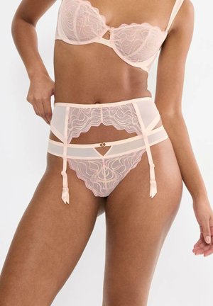 Kobieta w jasnoróżowym koronkowym komplecie bielizny składającym się z biustonosza, pasa do pończoch i dopasowanych majtek na białym tle.