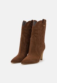 Ruske suede ankle boots z ostrimi nosi, dekorativnim šivanjem, ukrivljenim vrhom v zahodnem slogu in zmernim peto. Podrobnost z zlatim logotipom.