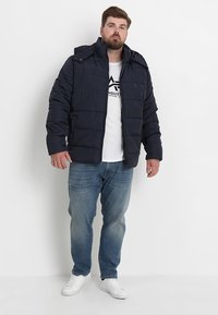 Marineblå dunjakke med quiltet design, full lynlås og fast hætte, båret over en hvid T-shirt og parret med lyseblå jeans og hvide sneakers.