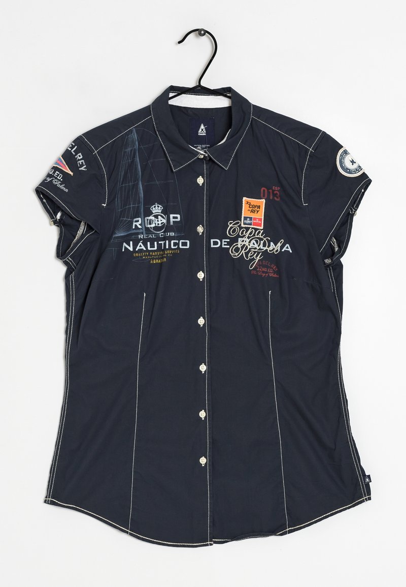 Chemise bleu marine à manches courtes boutonnée avec surpiqûres blanches, logos, texte et écussons à thème nautique, suspendue sur un cintre noir.