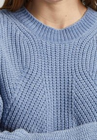 Suéter de punto azul claro con un cuello redondo acanalado, que presenta patrones texturizados y detalles en relieve a lo largo de la tela.