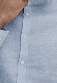 Camicia in lino azzurro chiaro con colletto button-down, caratterizzata da bottoni bianchi e un piccolo logo sul lato sinistro vicino al fondo.