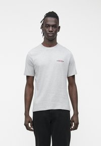 Szürke pamut rövid ujjú póló, a bal mellrészen egy kis piros és fekete "GUESS JEANS" logóval. Laza szabású, kerek nyakkivágással.