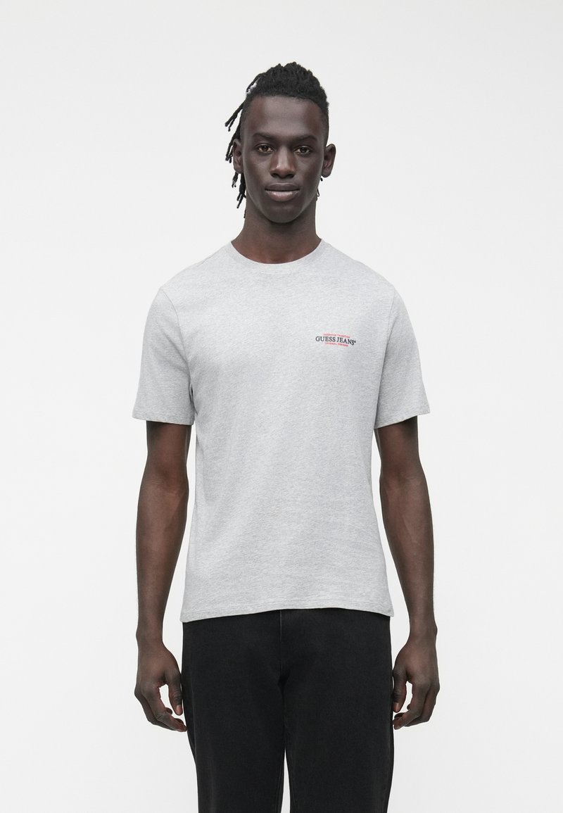 Szürke pamut rövid ujjú póló, a bal mellrészen egy kis piros és fekete "GUESS JEANS" logóval. Laza szabású, kerek nyakkivágással.