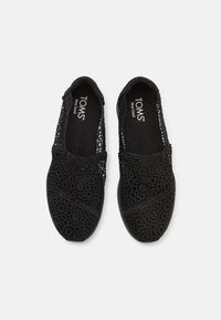 Musta pitsinen liukumekanavuori, jossa on monimutkainen kukkakuosi, pehmeä kangaspäällyste ja pyöristetty kärki. TOMS-logo näkyy sisäpohjassa.