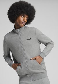 Šedá zipsová mikina s vysokým golierom, ktorá má bočné vrecká a čierne logo Puma na hrudi. Mäkká tkanina, predĺžené rukávy, minimalistický dizajn.