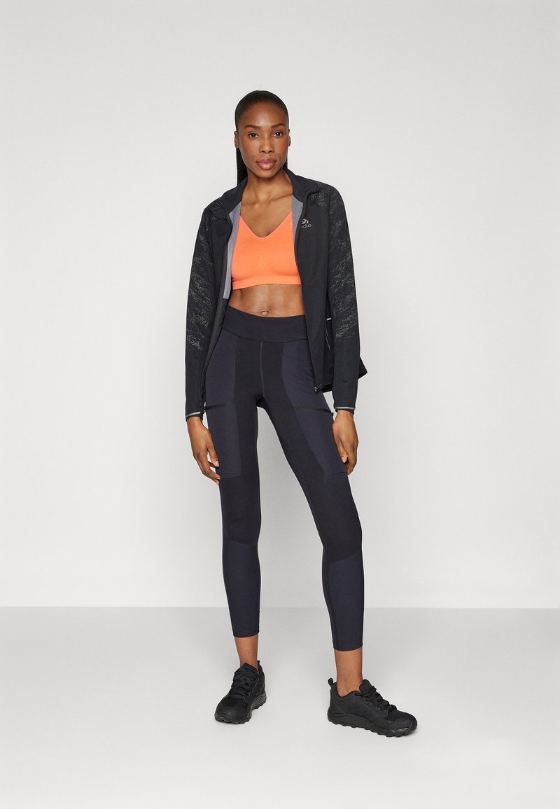 Schwarze Sportjacke mit strukturierten Ärmeln, orangefarbener Sport-BH und schwarze Leggings mit Paneelakzenten, kombiniert mit schwarzen Turnschuhen.