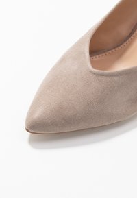 Beige mockaskinn spetsig tå platt sko med en låg vamp och inredning i tanläder på en vit bakgrund.