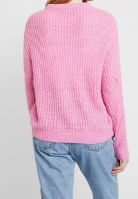 Flauschiger rosa Strickpullover mit strukturiertem Muster, geripptem Saum und überschnittenen Schultern, kombiniert mit hellblauen Jeans.