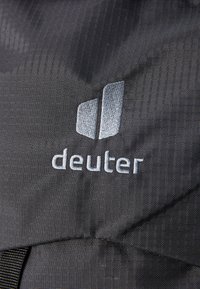 Fekete szövet, textúrált mintával, amelyen a világosszürke "deuter" logó hímezve található, egy sima területen elhelyezve.