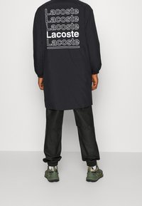 Černá bunda s dlouhými rukávy, která na zadní straně obsahuje opakující se bílé logo "Lacoste". V kombinaci s černými kalhotami a zelenými teniskami.