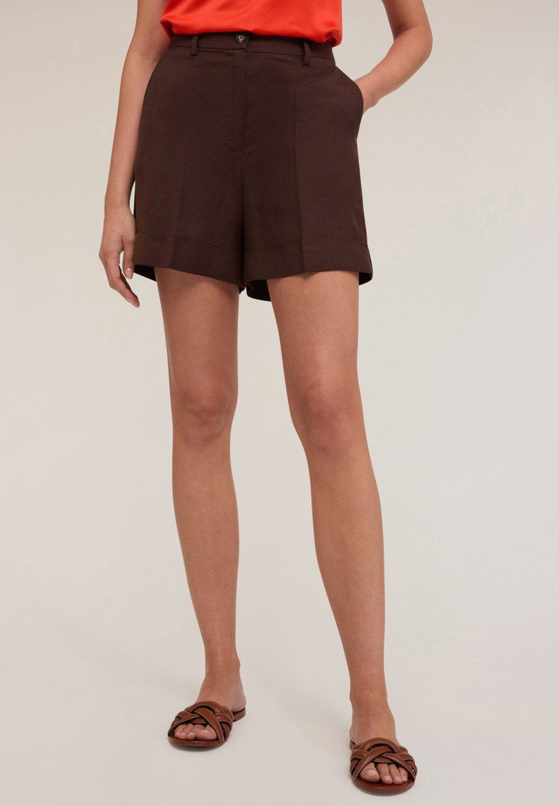Pantaloni corti in lino marrone con tasche laterali, orlo arrotolato e chiusura a bottone. Abbinati a sandali marroni con cinturini.