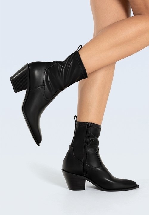 Botas de cuero negro hasta el tobillo con un diseño caído, punta afilada y un tacón cuadrado cónico. Presentan un zipper lateral y un detalle texturizado en el tacón.