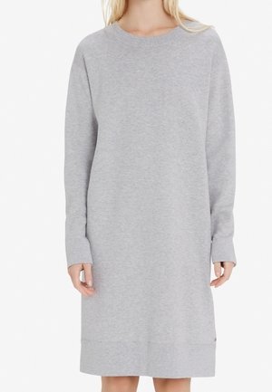 Vrouw draagt een lichtgrijze, knielange sweatshirtjurk met lange mouwen, een geribbelde ronde halslijn en manchetten.
