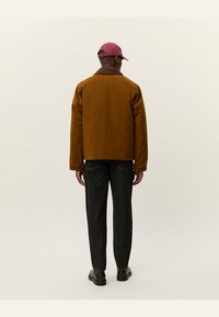 Giacca marrone realizzata in un materiale resistente, con colletto classico, abbinata a jeans scuri e un berretto rosa, vista di spalle.