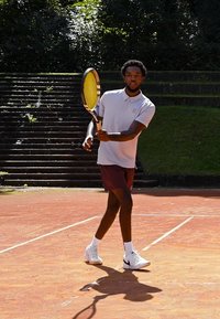 Tennis spiller i en lysegrå polo og mørke shorts, som holder et gult tennisketsjer, på en grusbane med stentrin i baggrunden.