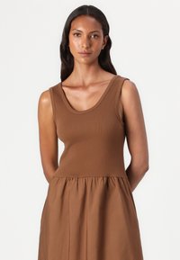 Brun ärmlös klänning med en figursydd bodice och en rynkad kjol. Tillverkad av mjukt tyg har den en slät yta och en rundad halsringning.
