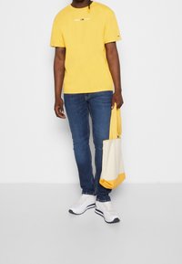 Yellow cotton t-shirt with embroidered logo, blue jeans, white sneakers, and a cream bag with yellow accents, held by a person.