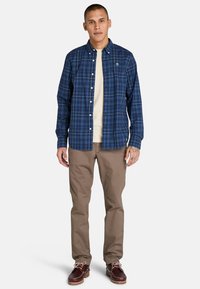 Modrá kostkovaná košile na knoflíky s logem, doplněná béžovými kalhotami a hnědými lodními botami. Košile má límec a bílé detaily knoflíků.