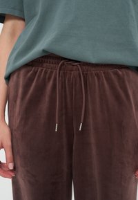 Gros plan d'une personne portant un pantalon en velours marron avec un cordon de serrage et un t-shirt vert ample, les mains détendues sur les côtés.