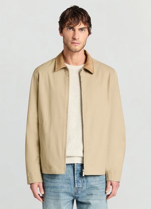 Veste beige avec un col marron clair, dotée d'une fermeture éclair à l'avant et de manches longues. Portée sur un pull léger et un jean bleu, présentant une texture lisse.