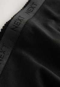 Abbigliamento in tessuto nero con una texture liscia e un bordo decorativo a scallop lungo il cinturino, con il logo "NEXT" in rilievo.