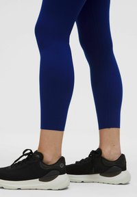 Alsó lábak sötétkék leggingsben és fekete sportcipőben, fehér talppal, egy sima világos háttér előtt állnak.