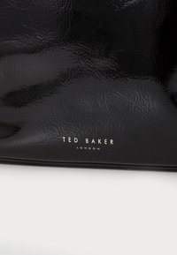 Bolso de mano de charol negro con textura grabada y detalle del logo en oro "Ted Baker London" en la parte frontal.