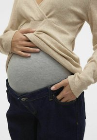 Bežový ovitni pulover preko sive pasne banda, roke dotikajo trebuha, v kombinaciji s temno modrimi jeans hlačami z rumenim šivom.